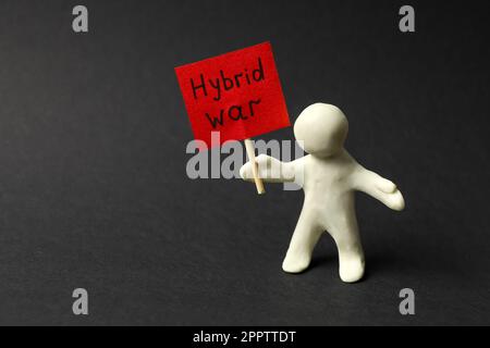 Figura umana fatta di plastilina bianca che tiene segno con le parole guerra ibrida su sfondo nero Foto Stock