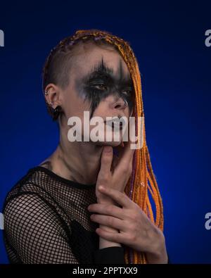 Ritratto cinematografico di una bella giovane donna con capelli color arancione e make-up nero horror scenico dipinto sul viso. Studio girato in blu Foto Stock