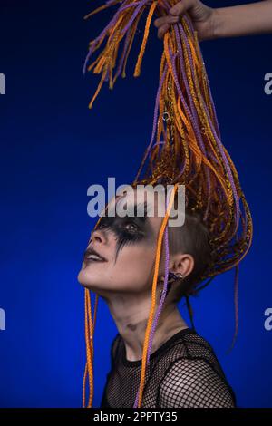 Giovane donna con acconciature color arancio e hairdo nero horror scena make up dipinto sul viso. Studio ha girato un ritratto spettacolare su sfondo blu Foto Stock
