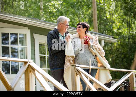 Coppia matura in piedi sul portico e bere caffè caldo da tazza. Sorridono e ridono, avvolti in una coperta. Coppia anziana felice abbracciando Foto Stock