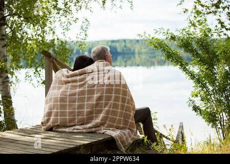 Coppia matura seduta su gradini di legno e abbracciante. Sono avvolti in coperta, sorridenti e guardano alla bellissima vista - la foresta e il lago. Buon anziano Foto Stock