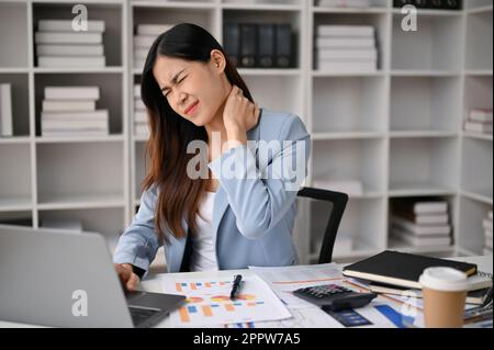 La donna d'affari asiatica, troppo lavorata e stanca, si siede alla scrivania del suo ufficio strofinando il collo, soffrendo di dolore al collo. Concetto di sindrome dell'ufficio Foto Stock