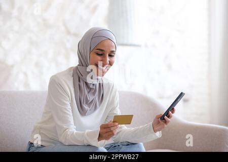 Giovane bella donna asiatica in hijab seduta sul divano a casa, donna musulmana tenendo la carta di credito e il telefono in mano, felicemente shopping online nel negozio online. Foto Stock