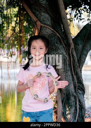 Bambina asiatica giocare il tamburello, nel giardino sotto il grande albero Foto Stock