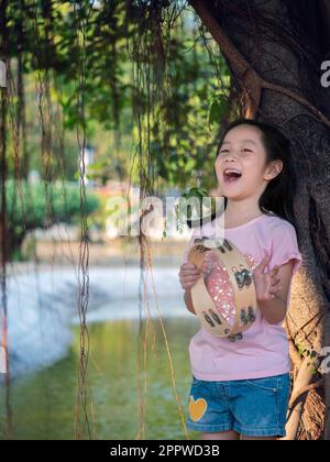 Bambina asiatica giocare il tamburello, nel giardino sotto il grande albero Foto Stock