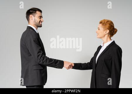 vista laterale di gioiosi partner commerciali in blazer neri che scuotono le mani isolate su grigio, immagine stock Foto Stock