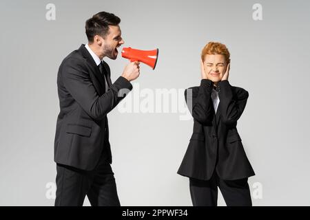 redhead manager con occhi chiusi che coprono le orecchie con le mani vicino al boss urlando in megafone isolato su grigio, immagine stock Foto Stock