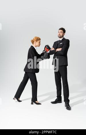 redhead businesswoman boxing vicino sorridente e scettico uomo d'affari in piedi con braccia piegate su sfondo grigio, immagine di scorta Foto Stock