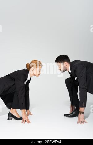 vista laterale di una donna d'affari rossa e di un uomo d'affari bearded in piedi in posizione di partenza bassa e guardando l'un l'altro su sfondo grigio, immagine di scorta Foto Stock