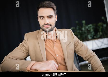 uomo d'affari positivo ed elegante in blazer beige seduto in ufficio e guardando la fotocamera, immagine stock Foto Stock