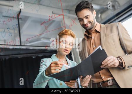 businesswoman in occhiali guardando documenti vicino sorridente collega e lavagna in vetro in ufficio, immagine stock Foto Stock