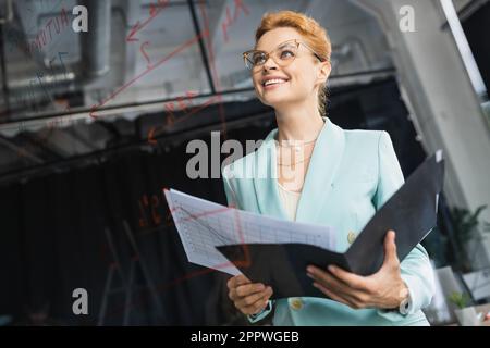 vista ad angolo basso di una donna d'affari soddisfatta che tiene la cartella con i documenti mentre guarda l'ufficio di bordo di vetro, immagine di scorta Foto Stock
