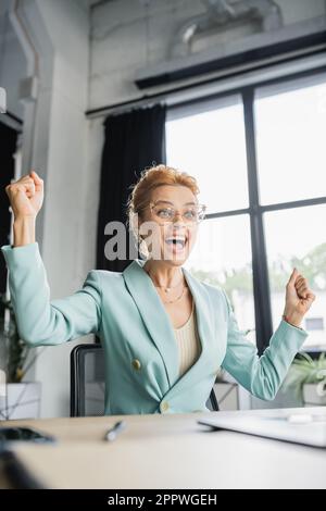 overjoyed redhead businesswoman in eyeglasses mostrando il gesto di vittoria e urlando in ufficio, immagine di riserva Foto Stock