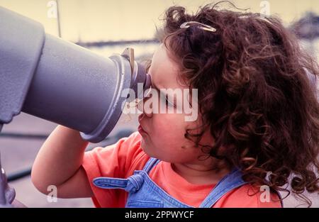 Primo piano di un bambino con capelli ricci marroni che guardano attraverso il mirino di un telescopio a gettoni sul mare. Foto Stock