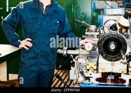 Un uomo che lavora in un accappatoio, tute si trova accanto ad un tornio industriale per tagliare, ruotare coltelli da metalli, legno e altri materiali, tornire, makin Foto Stock