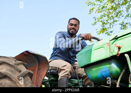 Ritratto di giovane agricoltore maschio che guida trattore verde con cielo limpido sullo sfondo nelle giornate di sole Foto Stock