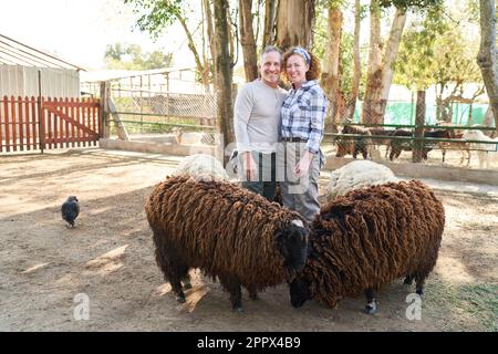 Ritratto di sorridente coppia contadina matura in piedi con pecore contro gli alberi in fattoria Foto Stock