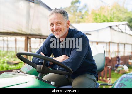 Ritratto di un contadino maschio maturo sorridente seduto sul trattore con serra sullo sfondo Foto Stock