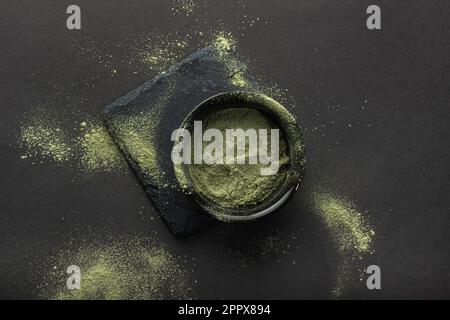 Polvere verde in una piccola ciotola di ceramica, su un cavalletto di pietra, su fondo nero. Lo sfondo intorno è spruzzato con questa polvere. Super cibo o ma Foto Stock