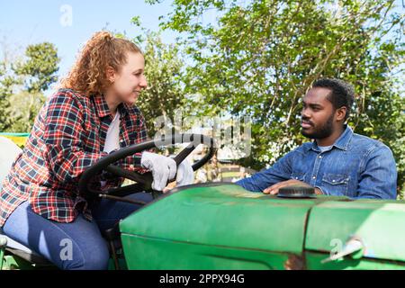 Sorridenti contadini multiculturali di sesso maschile e femminile che discutono mentre si siedono sul trattore in azienda nelle giornate di sole Foto Stock