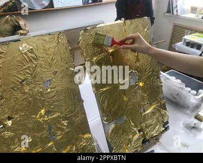 La mano dell'artista dipinge con un pennello utilizzando oro giallo brillante foglia d'oro metallico per scopi artistici e creativi su tela. Il backg Foto Stock