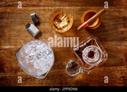 Vista di un vetro di cristallo e di un decanter pieno di whisky dorato, secchio di ghiaccio, sigaro e accendino, girato dall'alto su uno sfondo di legno scuro Foto Stock