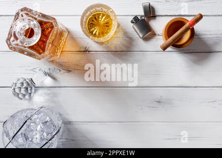 Vista di un vetro di cristallo e di un decanter pieno di whisky dorato, secchio di ghiaccio, sigaro e accendino a benzina, girato dall'alto su un cinghiale di legno bianco afflitto Foto Stock