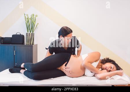 Vista laterale dell'osteopata giovane che tratta la paziente in gravidanza guardando la telecamera e sdraiata sul tavolo mentre tratta il bambino nella pancia per la posizione corretta durante la s. Foto Stock