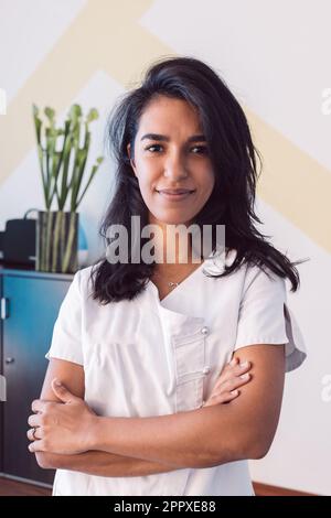Donna positiva che indossa uniforme medica bianca e braccia incrociate mentre guarda la fotocamera in ufficio Foto Stock