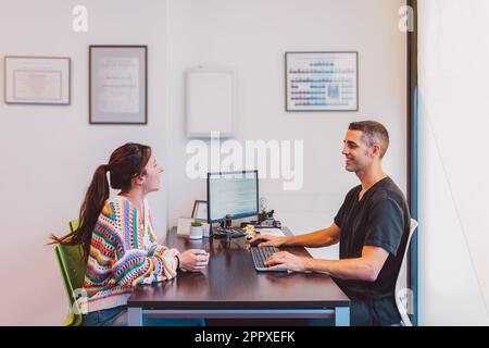 Vista laterale di un medico maschio positivo in maglietta nera seduta a tavolo con il computer e parlando con la paziente femminile mentre si discutono i risultati del trattamento Foto Stock