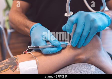 Medico irriconoscibile in guanti di lattice che trattano il paziente con apparecchiature a ultrasuoni durante l'esame della gamba durante l'appuntamento in ospedale contemporaneo Foto Stock