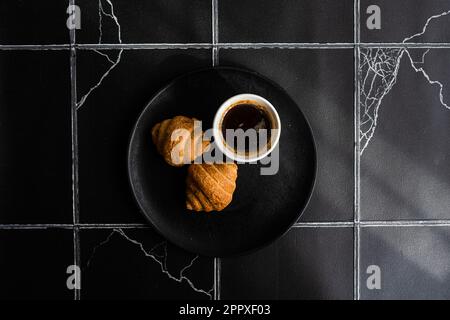 Vista dall'alto di un caffè nero e croissant appena sfornati su un tavolo di cemento nero Foto Stock
