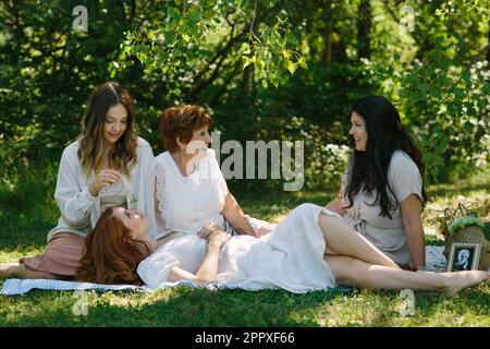 Corpo pieno di allegre femmine multi generazionali sorridenti e guardandosi mentre riposano sulla coperta da picnic e parlano insieme nel parco verde Foto Stock
