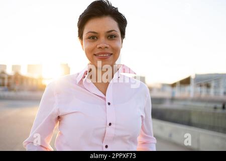 Donna afro-americana positiva in camicia rosa con sorriso toothy e capelli corti guardando la macchina fotografica mentre si trova su sfondo sfocato della città Foto Stock