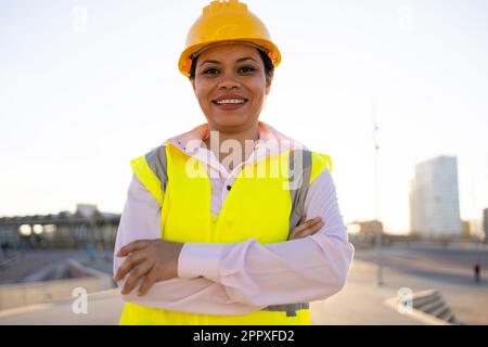 Giovane ingegnere di etnia positiva in hardhat e corpetto sorridendo e guardando la macchina fotografica mentre si è in piedi sulla strada della città contro il cielo nuvoloso Foto Stock