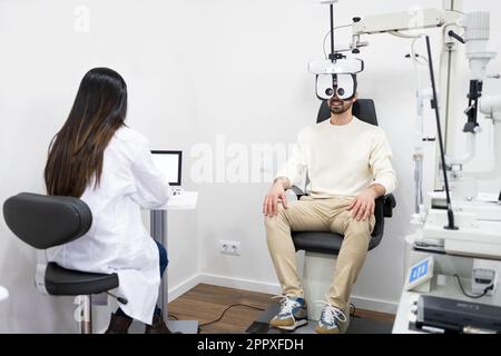 Corpo pieno di giovane cliente sorridente maschio seduto sulla sedia vicino al medico dell'occhio e guardando attraverso il foropter ottico durante l'esame dell'occhio in clinica Foto Stock