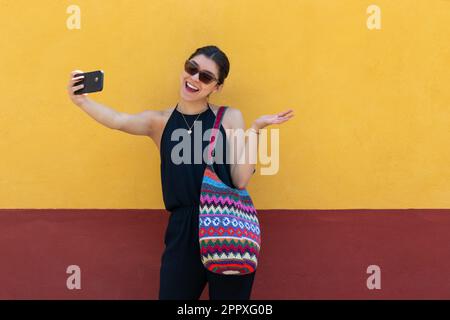 Ragazza positiva in abiti alla moda e occhiali da sole sorridente e scattando un selfie mentre si è in piedi contro la parete gialla e parlando su smartphone all'aperto Foto Stock