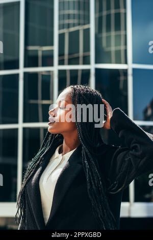 Giovane ragazza afroamericana con trecce afroamericane e giacca nera in piedi sulla strada cittadina con mano sulla testa e occhi chiusi Foto Stock