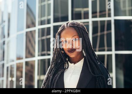 Giovane donna nera con trecce Afro che guarda la macchina fotografica mentre si trova vicino a una parete di vetro sulla strada cittadina Foto Stock