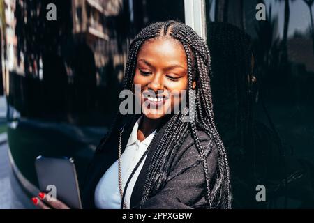 Donna afro-americana positiva in abiti casual e con trecce afro sorridendo e navigando il telefono cellulare mentre si trova in piedi in città strada il giorno di sole Foto Stock