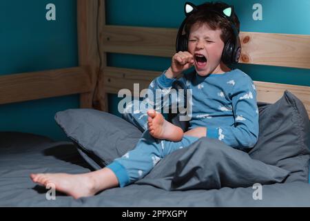Felice bambino in abito da notte urlare con eccitazione con gli occhi e pugni chiusi mentre si ascolta la musica in cuffie wireless e appoggiarsi su tavola di legno Foto Stock
