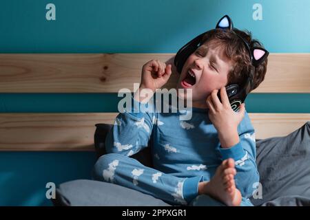 Felice bambino in abito da notte urlare con eccitazione con gli occhi e pugni chiusi mentre si ascolta la musica in cuffie wireless e appoggiarsi su tavola di legno Foto Stock