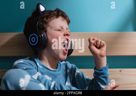 Felice bambino in abito da notte urlare con eccitazione con gli occhi e pugni chiusi mentre si ascolta la musica in cuffie wireless e appoggiarsi su tavola di legno Foto Stock