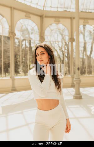 Affascinante giovane donna in bianco con lunghi capelli castani guardando la macchina fotografica mentre si è in piedi in Palazzo di vetro nel Parco del Buen Retiro a Madrid Foto Stock