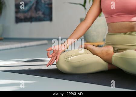 Donna irriconoscibile in abbigliamento sportivo meditare in posa Padmasana mentre si pratica yoga con le mani mudra Gyan sul tappeto durante la sessione in studio luce Foto Stock