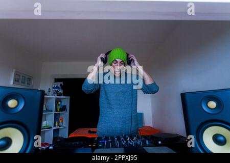 Uomo adulto concentrato in cappello verde e maglione blu in piedi vicino alla console di mixaggio DJ e indossare le cuffie nel soggiorno Foto Stock