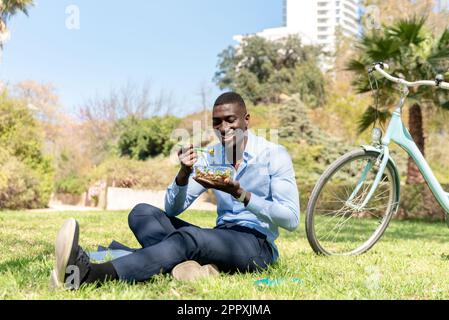 Un uomo d'affari afro-americano sorridente che indossa abiti di classe seduti sull'erba verde e gustando cibo sano mentre pranza vicino al bic Foto Stock