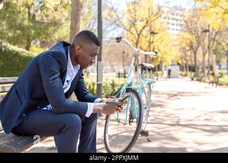 Vista laterale del manager afro-americano maschile in abito di classe seduto su una panca di legno vicino alla bicicletta nel parco e navigare sul telefono cellulare durante il giorno Foto Stock