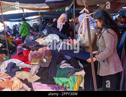 Sousse, Tunisia, 15 gennaio 2023: Giovane tunisina dai capelli neri ben vestita controlla l'offerta tessile sul tavolo da presa vestiti nel mercato Foto Stock