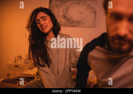 Donna insoddisfatta seduta vicino boyfriend sfocato in camera da letto di notte, immagine di scorta Foto Stock